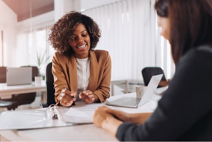 A woman in a business setting smiles and gestures while discussing paperwork with a colleague across a desk, with a laptop and documents nearby.