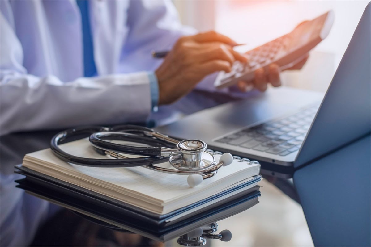 Hands of a healthcare professional use a calculator beside a laptop, with a stethoscope resting on a notebook in the foreground, suggesting medical billing or administrative work.