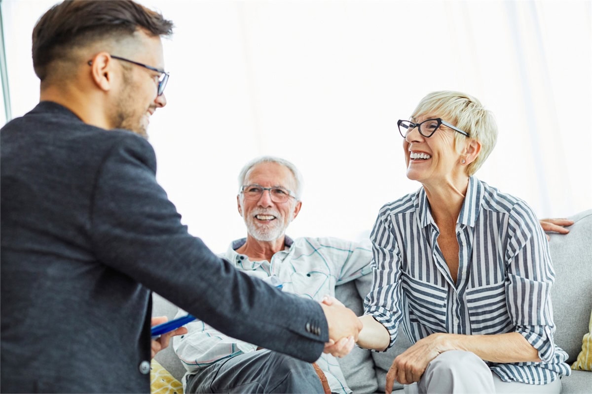 A professional shakes hands with a smiling older woman while an older man sits beside her, suggesting a friendly meeting or consultation in a home setting.