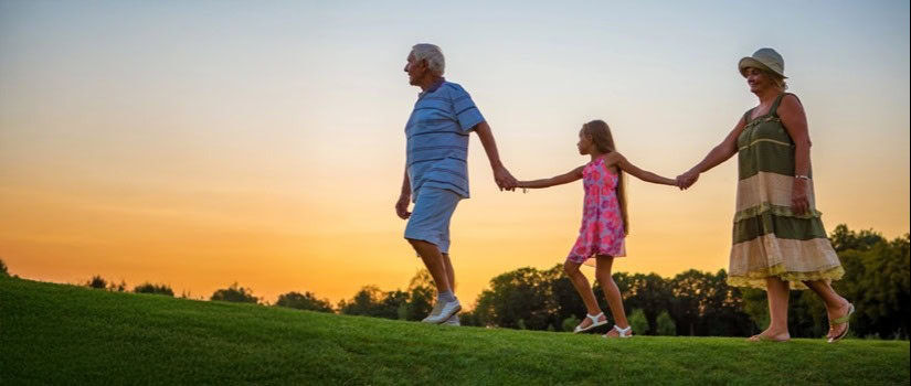 An older couple walks hand in hand with a young girl across a grassy hill at sunset, enjoying time together outdoors.