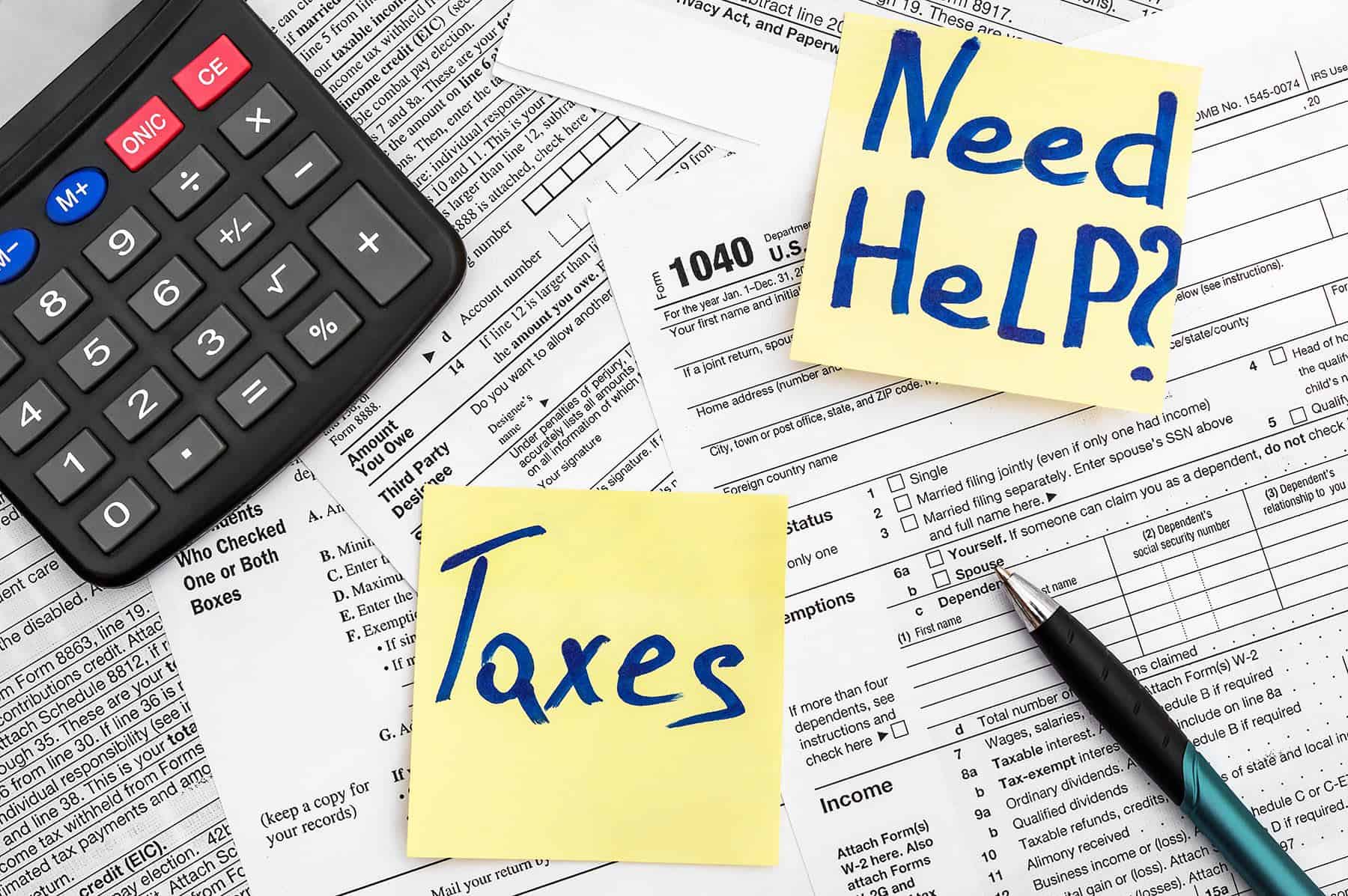 A calculator, tax forms, and sticky notes reading "Taxes" and "Need Help?" with a pen nearby